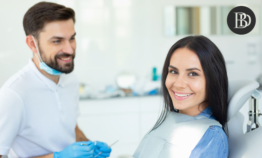 A young female patient sits on the dentist's chair and smiles at the camera.